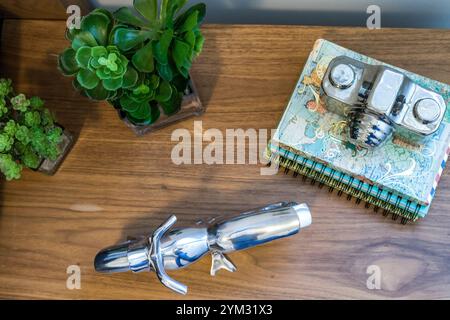 Ein silbernes Objekt sitzt auf einem Holztisch neben einem Notizbuch und einer Topfpflanze. Das silberne Objekt ist eine Kamera, und das Notizbuch ist für eine Seite mit einem geöffnet Stockfoto