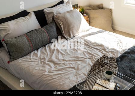 Ein Bett mit einer weißen Decke und schwarz-weißen Kissen. Ein Tablett mit einer Tasse und einem Buch liegt auf dem Bett Stockfoto