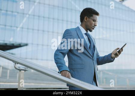 Geschäftsmann in blauem Anzug, der tagsüber auf einer Treppe vor dem modernen Glasgebäude sein Smartphone überprüft. Hintergrund ist eine architektonische Struktur mit Metallgeländern Stockfoto