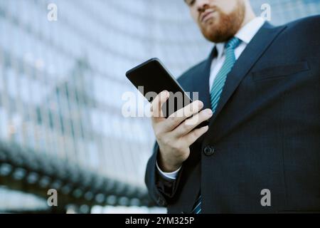 Mann im formellen Anzug, der ein Smartphone hält, während er vor einem modernen Glasgebäude steht. Er sieht konzentriert aus, während er mit dem Gerät interagiert Stockfoto