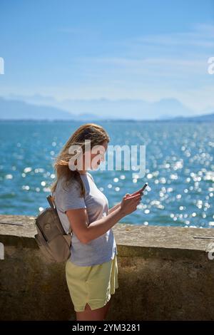 Frau, die an einem sonnigen Tag Fotos an einem glitzernden See mit Panoramablick auf die Berge macht. Frau, die an einem sonnigen Tag malerische Fotos an einem glitzernden See macht Stockfoto