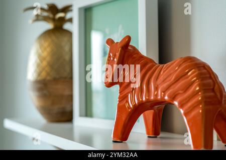 Eine Zebrafigur sitzt auf einem Regal neben einer Ananas und einem gerahmten Bild. Das Konzept von Wärme und Gemütlichkeit, mit orangefarbenem Zebra, das einen Farbakzent verleiht Stockfoto