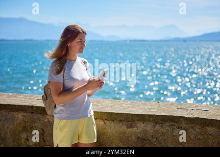 Frau, die an einem sonnigen Tag Fotos an einem glitzernden See mit Panoramablick auf die Berge macht. Frau, die an einem sonnigen Tag malerische Fotos an einem glitzernden See macht Stockfoto