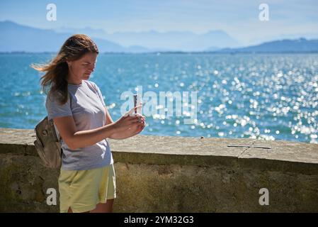 Frau macht an einem sonnigen Tag mit Blick auf die majestätischen Berge malerische Fotos an einem glitzernden See. Idyllische Lage am Seeufer bietet natürliche Schönheit und O Stockfoto