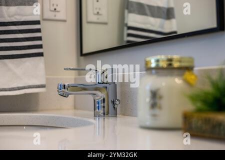 Ein Waschbecken im Badezimmer mit silbernem Wasserhahn und einem Glas duftender Kerze auf der Theke. Die Kerze wird neben eine kleine Pflanze gestellt, was einen Hauch von Grün verleiht Stockfoto