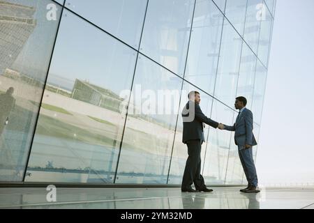 Zwei Geschäftsleute schütteln sich die Hände vor einem großen Glasgebäude, das Himmel und Landschaft reflektiert. Beide Personen tragen formelle Anzüge, die einen Geschäftsvertrag besiegeln Stockfoto