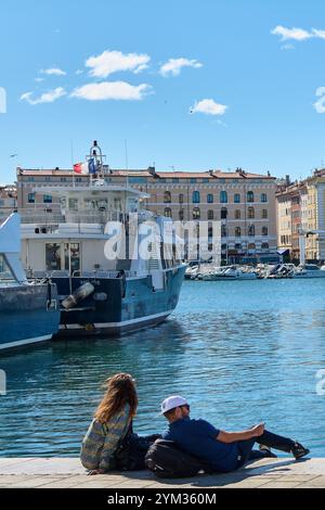 Marseille. Frankreich - 20. November 2024: Junges Paar, das sich im Hafen von Marseille entspannt, mit Panoramablick auf Yachten und Gebäude im Hintergrund, Stockfoto