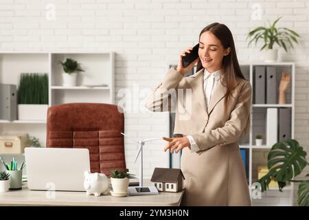 Weibliche Ingenieurin, die mit dem Handy spricht, mit einem Windkraftanlagenmodell auf dem Tisch im Büro Stockfoto