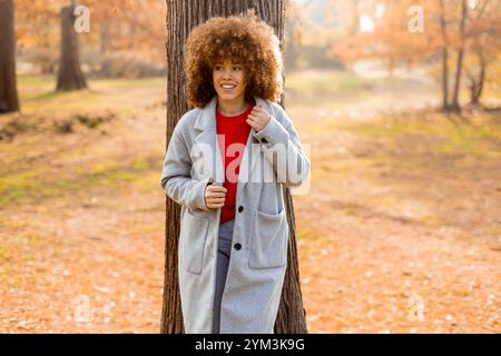 Eine junge Frau mit lockigen Haaren lehnt sich an einen Baum, trägt einen warmen Pullover, lächelt fröhlich im goldenen Herbstlaub und umhergefallenen Blättern Stockfoto