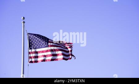 Eine verwitterte amerikanische Flagge weht in einer sanften Brise gegen einen blassblauen Himmel. Die Flagge ist an einem hohen Fahnenmast mit einem einfachen Finial befestigt. Stockfoto