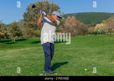 Der ältere Golfer macht mit seinem Schläger einen langen Schuss und zielt während des Trainings oder eines Spiels auf dem Golfplatz auf das Loch auf dem Grün Stockfoto