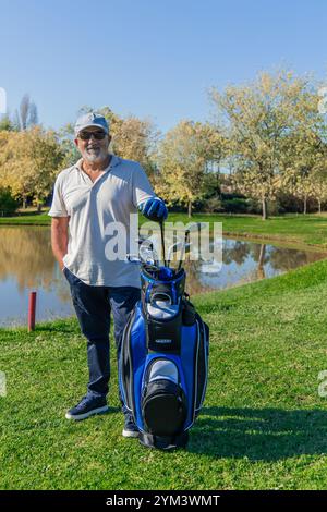 Vertikaler älterer Golfer mit Sonnenbrille und Kappe lehnt sich an seinen Golfbeutel neben dem ruhigen See auf dem Golfplatz und genießt einen Moment der Entspannung Stockfoto