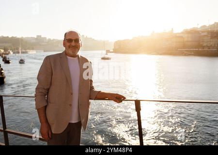 Mann in einer leichten Jacke, der den Sonnenuntergang am Ufer in einem geschäftigen Hafen genießt Stockfoto