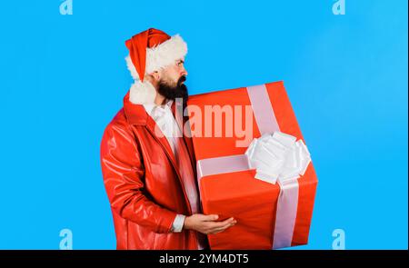 Weihnachtseinkäufe. Ernsthafter bärtiger Mann mit Weihnachtsmütze mit Weihnachtsgeschenk. Neujahrsgeschenk. Hübscher Mann im roten Mantel mit Weihnachtsgeschenkkasten. Fröhlich Stockfoto