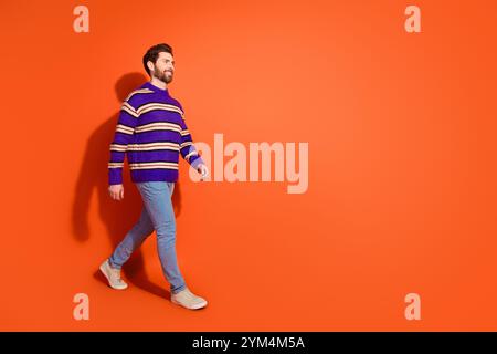 Stilvoller junger Mann in gestreiftem Pullover, der vor leuchtendem orangefarbenem Hintergrund läuft und moderne Mode zeigt Stockfoto