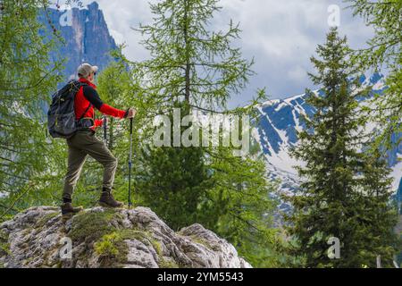 Ein Wanderer steht auf einem großen Felsen und genießt die atemberaubende Berglandschaft umgeben von hohen Bäumen. Wolken füllen den Himmel und schaffen eine dramatische Kulisse. Stockfoto