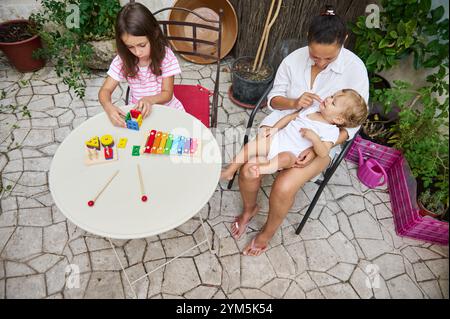 Eine Mutter hält ihr Baby liebevoll in der Hand, während ihre Tochter mit bunten Lernspielzeugen im Garten spielt. Stockfoto