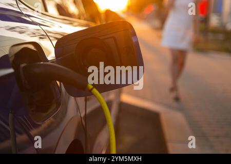Schließen Sie das Aufladen eines Elektroautos, während eine Frau in einem weißen Kleid bei Sonnenuntergang neben ihm läuft. Stockfoto