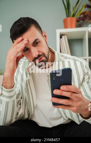Besorgter junger Mann, der ein Smartphone hält, sich gestresst und frustriert fühlt, wenn er schlechte Nachrichten liest oder ein Problem hat, drinnen in einer ungezwungenen Umgebung sitzt, Besorgnis und Nachsicht zum Ausdruck bringt. Hochwertige Fotos Stockfoto