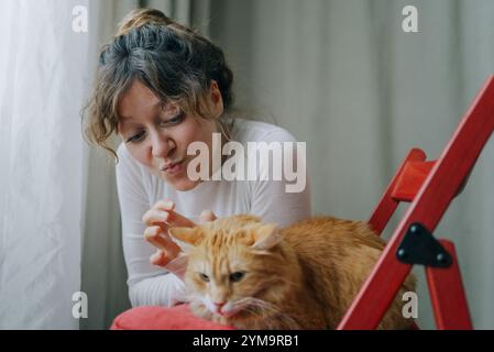 Eine junge Frau, die ein weißes Hemd trägt, macht ein lustiges Gesicht, während sie ihre Ingwerkatze streichelt, die auf einem roten Stuhl neben einem Fenster mit weißen Vorhängen sitzt Stockfoto