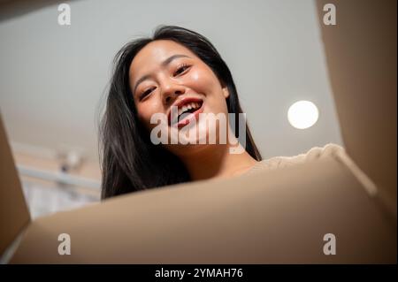 Eine fröhliche, aufgeregte asiatische Frau öffnet zu Hause eine Geschenkbox. Ansicht von der Innenseite des Kastens. Geburtstagsgeschenk, Weihnachtsgeschenk, Paketlieferung Stockfoto