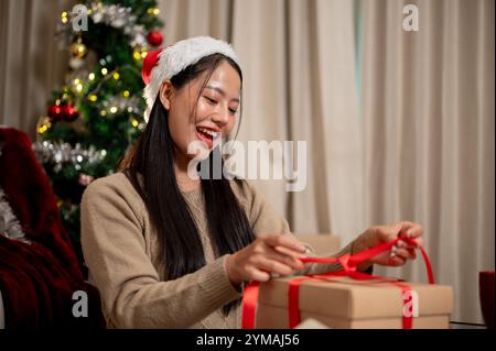 Eine attraktive, lächelnde asiatische Frau in gemütlicher Kleidung bereitet im Wohnzimmer Weihnachtsgeschenke vor und bindet ein rotes Band um eine Geschenkbox. Urlaub, Chri Stockfoto