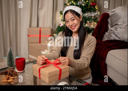 Eine charmante asiatische Frau bereitet im Wohnzimmer Weihnachtsgeschenke vor, indem sie vorsichtig ein rotes Band um eine Geschenkbox bindet und die Kamera anlächelt. Stockfoto