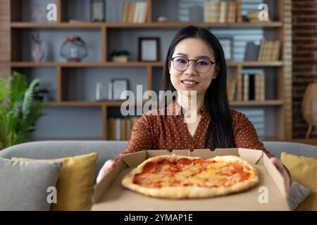 Asiatische Frau in braunem Hemd, die Pfefferoni-Pizza in Pappkarton hält und sie mit einem Lächeln im gemütlichen Wohnzimmer anbietet. Wohnanlage mit moderner Einrichtung, Bücherregalen, komfortabler Couch. Stockfoto