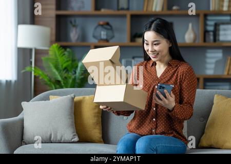 Asiatische Frau, die auf dem Sofa sitzt und das gelieferte Paket mit dem Smartphone in der Hand öffnet. Drückt die Begeisterung für Online-Shopping und den Empfang von Bestellungen zu Hause aus. Steht für ein freudiges Unboxing-Erlebnis. Stockfoto