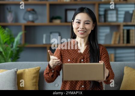 Asiatische Frau, die Pappkarton hält, auf dem Sofa sitzt und warm lächelt. Perfekte Darstellung von Lieferung nach Hause, Produkt-Auspacken, Geschenk geben, gemütliche Umgebung, warme Einladung. Stockfoto