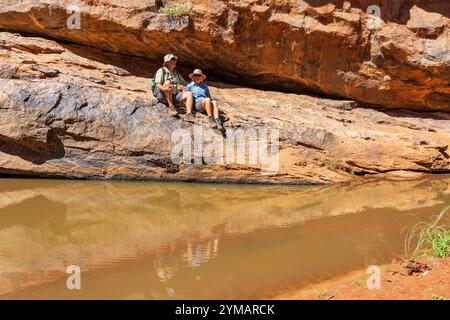 Ein paar Wanderer sitzen auf einem Felsvorsprung über einem Wasserloch im Mutawintji National Park im Outback New South Wales, Australien. Stockfoto