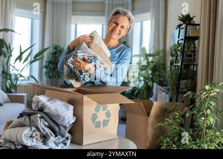 Grauhaarige, reife, lächelnde Frau verpacken Kleidung für das Recycling in den Karton zu Hause Stockfoto