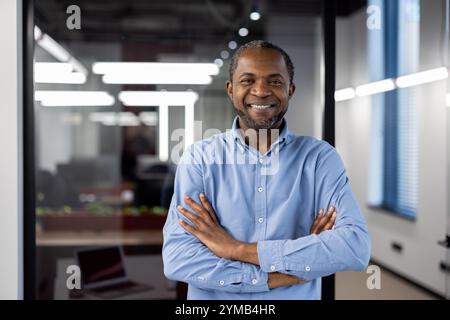 Selbstbewusster afroamerikanischer Geschäftsmann, der in einem modernen Büro steht. Mann mit blauem Hemd, lächelt mit überkreuzten Armen. Unternehmensumgebung mit heller Beleuchtung und Glasabtrennungen. Stockfoto