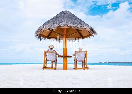 Ein junges verheiratetes Paar sitzt unter einem Strohschirm auf Holzstühlen am Strand. Die Aussicht von hinten. Ein Mann und eine Frau entspannen und ruhen sich bei Th aus Stockfoto