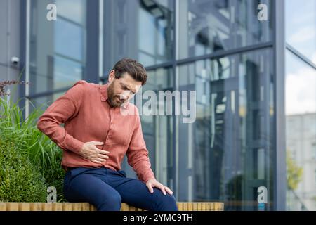Reifer Geschäftsmann mit Magenschmerzen sitzt draußen in einem modernen Geschäftsviertel. Er hält den Bauch, zeigt Unbehagen und Stress, symbolisiert gesundheitliche Probleme, sucht Linderung. Stockfoto
