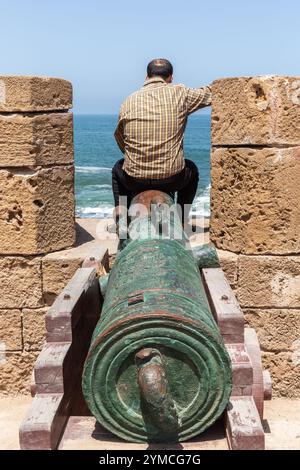 Auf einer alten Kanone in der Festung Essaouira steht ein Mann, der das Meer betrachtet Stockfoto