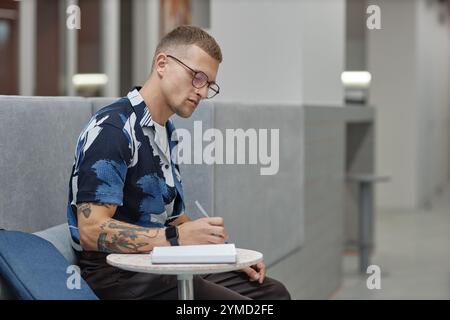 Seitenansicht eines jungen männlichen Geschäftsarbeiters, der sich auf das Schreiben in einem Notizbuch konzentriert und am Tisch sitzt, im Gemeinschaftsbereich des modernen Bürogebäudes, Kopierraum Stockfoto