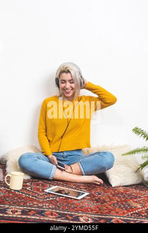 Kaukasische blonde Frau in einem gelben Pullover, lächelt und sitzt im Kreuz, trägt Kopfhörer, hält ein Telefon, genießt Musik drinnen. Stockfoto