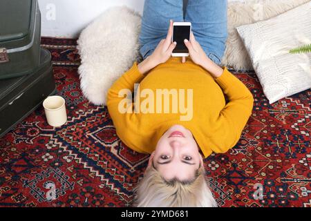 Weiße Frau mit blonden Haaren, gelbem Pullover und Jeans, die auf einem gemusterten Teppich liegt und ein Smartphone hält, entspannt zu Hause. Die Einrichtung umfasst Koffer A Stockfoto
