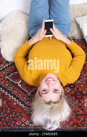 Weiße Frau mit blonden Haaren, gelbem Pullover und Jeans, die auf einem gemusterten Teppich liegt und ein Smartphone hält, entspannt zu Hause. Die Einrichtung umfasst Koffer A Stockfoto