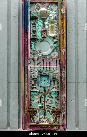 Eine Zusammenstellung aus der Wandinstallation „Memora di Kaya“ von TOPO 13. Alltägliche Haushaltsgegenstände – eine Porzellankanne, ein dekorativer Teller, ein Kerzenleuchter und ein Wecker – sind in einem alten Fensterrahmen auf Glasmosaiken angeordnet und bilden ein Stillleben, das das Leben des Haushalts darstellt. Willemstad, Curacao, Kòrsou Stockfoto