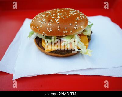 Ein Cheeseburger mit Salat und Käse wird auf eine Serviette auf einem roten Tablett gelegt. Das Brötchen ist braun und hat Sesamsamen. Der Burger wird auf einem roten Tablett serviert, was der Szene einen Farbakzent verleiht Stockfoto