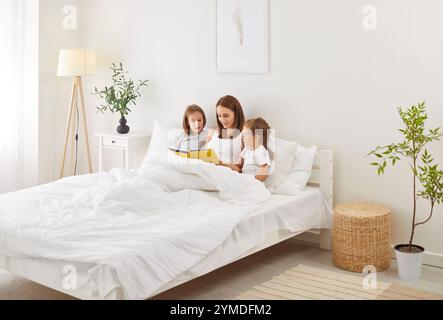 Mutter und zwei Kinder liegen im Bett unter der Decke, lesen zusammen Buch und genießen ein Familienwochenende. Stockfoto