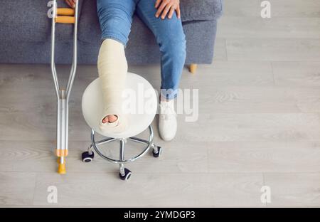 Nahaufnahme von gekrümmten Krücken und gebrochenem, bandagiertem Bein aus Gips auf dem Stuhl. Draufsicht Stockfoto