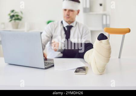 Behinderter Geschäftsmann, der am Laptop arbeitet und am Schreibtisch im Büro sitzt, mit bandagiertem Kopf und Krücken Stockfoto