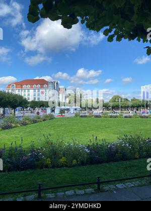 Ein großes Gebäude mit einem roten Dach befindet sich vor einem üppig grünen Park. Der Park ist voller Menschen, die den schönen Tag genießen. Polen, Sopot 28.08.2023 Stockfoto