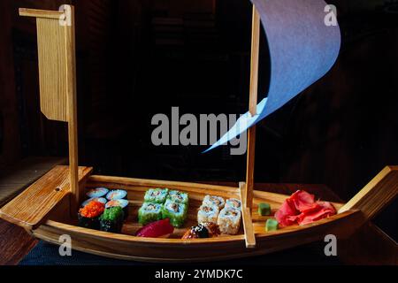 Ein Holzboot mit einem Segel ist mit verschiedenen Sushi-Rollen gefüllt. Das Boot wird auf einen Tisch gestellt und das Sushi ist so angeordnet, dass es wie ein echtes Boot aussieht Stockfoto