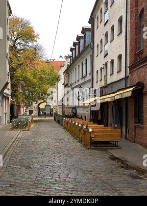 Eine kopfsteingepflasterte Straße mit Markisen und Tischen. Es gibt mehrere Bänke und Stühle. Die Straße ist von Bäumen gesäumt und hat Markisen über den Tischen. Lettland, Riga 08.10.2023 Stockfoto