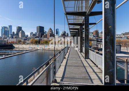 Ein schmaler Steg aus Holz und Metall führt den Betrachter in Richtung der beeindruckenden Skyline von Montreal, Kanada. Die Perspektive entlang der Brücke erzeugt eine starke Tiefe und lenkt den Blick auf die modernen und historischen Gebäude im Hintergrund. Auf der linken Seite spiegeln sich die Strukturen des Alten Hafens im ruhigen Wasser, während der blaue Himmel und das herbstliche Licht die Szenerie aufhellen. Die Mischung aus urbanem Charme, industrielle Details und architektonischer Vielfalt macht dieses Foto zu einem faszinierenden Einblick in das Herz von Montreal. *** Ein schmaler Woo Stockfoto