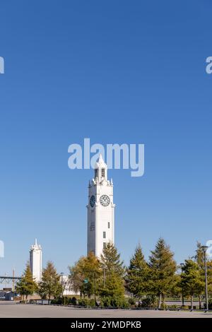 Der historische Uhrturm im Alten Hafen von Montreal, Kanada, erhebt sich majestätisch vor einem strahlend blauen Himmel. Der weiße Turm mit seiner eleganten Architektur und der markanten Uhr ist von gepflegten Bäumen und Grünflächen umgeben, die Bild eine ruhige und natürliche Atmosphäre verleihen. Im Hintergrund ist ein zweiter, kleinerer Turm zu erkennen, der die symmetrische Gestaltung des areals unterstreicht. Der Kontrast zwischen dem klaren Himmel, den grünen Bäumen und dem weißen Turm sorgt für eine harmonische und ausdrucksstarke Komposition. *** Der historische Glockenturm in der Alten Stockfoto